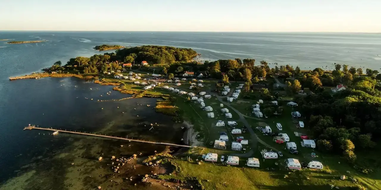 Vacker vy över Kronobergs camping med husvagnar och husbilar utspridda bland träd och vid vattnet. En brygga sträcker sig ut i det klara vattnet.