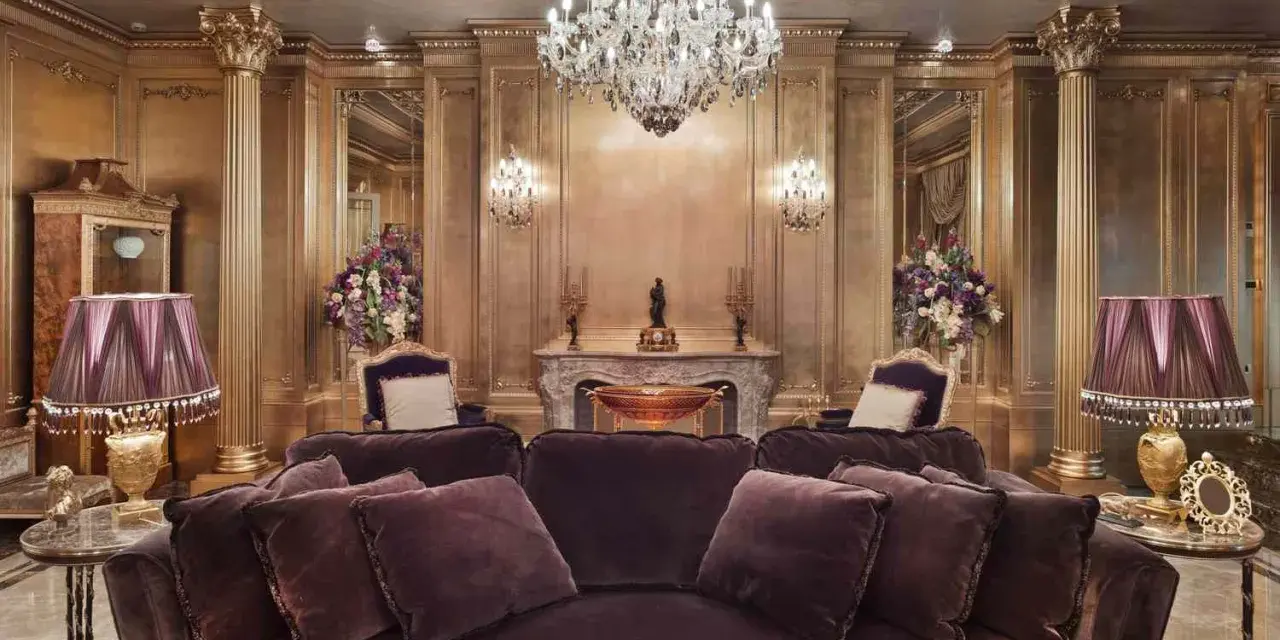 Salón lujoso con sofás de terciopelo morado, lámparas doradas y un candelabro de cristal. El techo y las paredes doradas evocan el estilo barroco español.