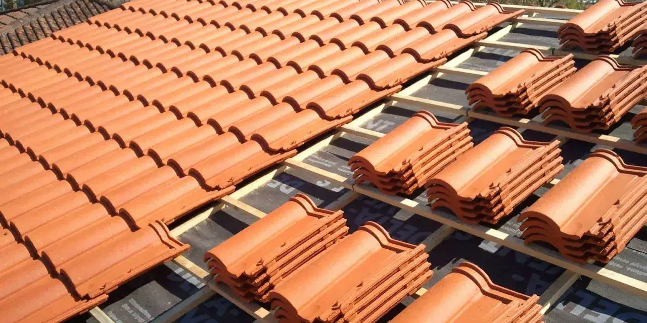 Vue d'un toit en cours de pose, révélant des rangées de tuiles mécaniques en terre cuite orange vif, empilées sur des lattes de bois.