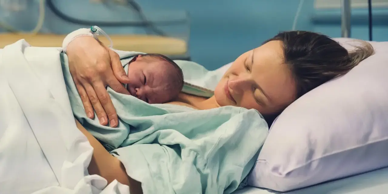 Eine Mutter hält ihr Neugeborenes im Arm, glücklich und erschöpft. Die Szene zeigt, wie lange nach Geburt im Krankenhaus die erste Bindung entsteht.