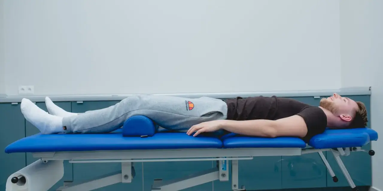 Mężczyzna leży na kozetce rehabilitacyjnej, z podparciem pod kolanami. Rozpoczęła się jego sis rehabilitacja.