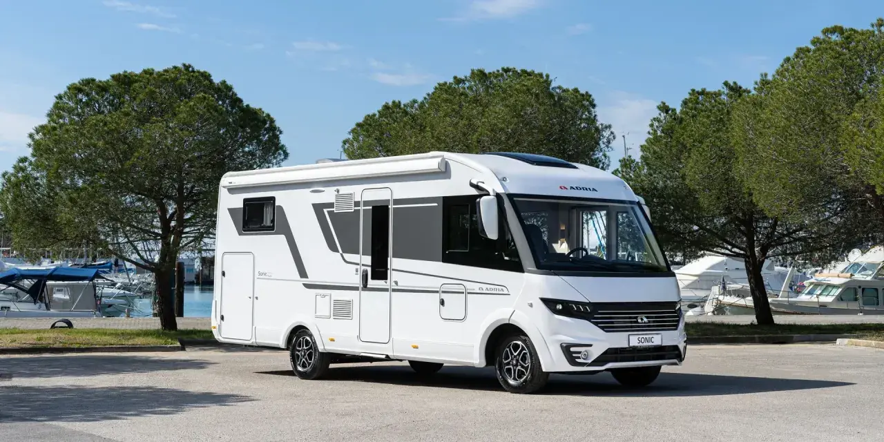 En vit Adria Sonic husbil, den lyxigaste husbilen, parkerad vid en marina med båtar och träd i bakgrunden.
