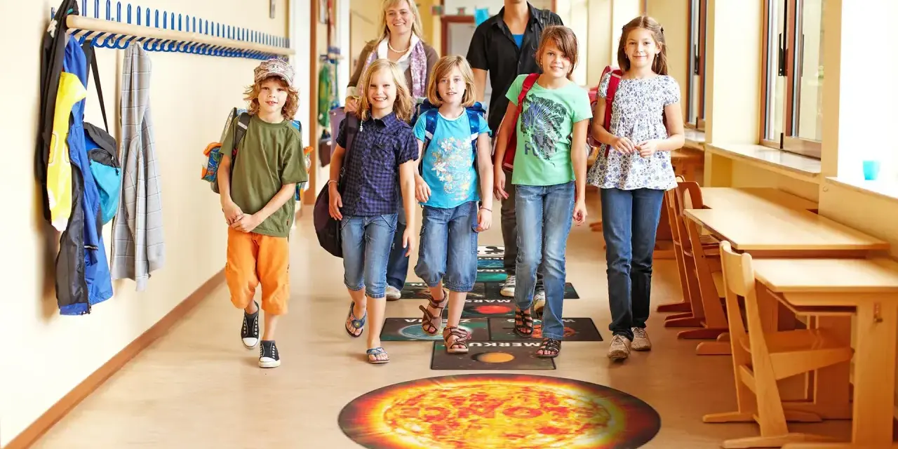 Dzieci i dorośli idą szkolnym korytarzem, stąpając po planszy z planetami. Fajne inicjatywy w szkole promują naukę przez zabawę.