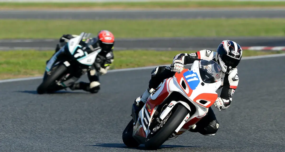 Dwóch motocyklistów w pełnym rynsztunku ściga się na torze. Jeden z nich, z numerem 11, pochyla się nisko w zakręcie, demonstrując emocje sportu motocyklowego.