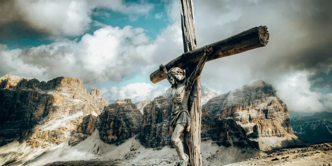 Jesus am Kreuz, eine hölzerne Figur, steht vor majestätischen Bergen unter einem dramatischen Himmel.