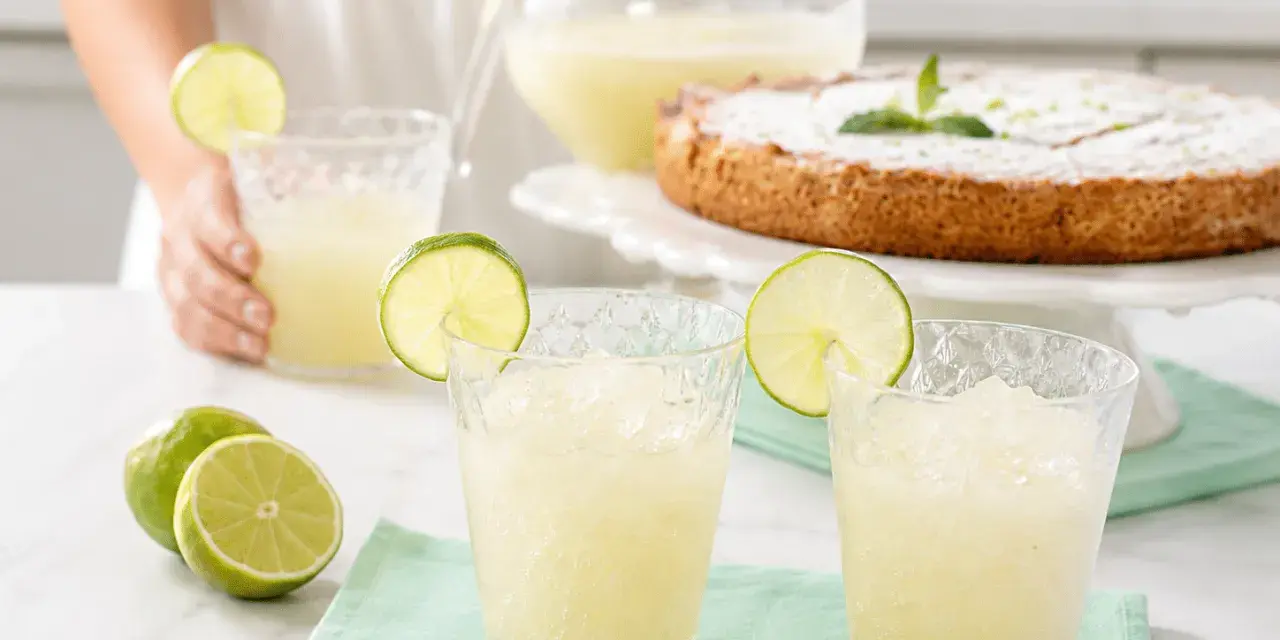 Dos vasos de granizado de limón refrescante, adornados con rodajas de lima, junto a limones frescos y un pastel.