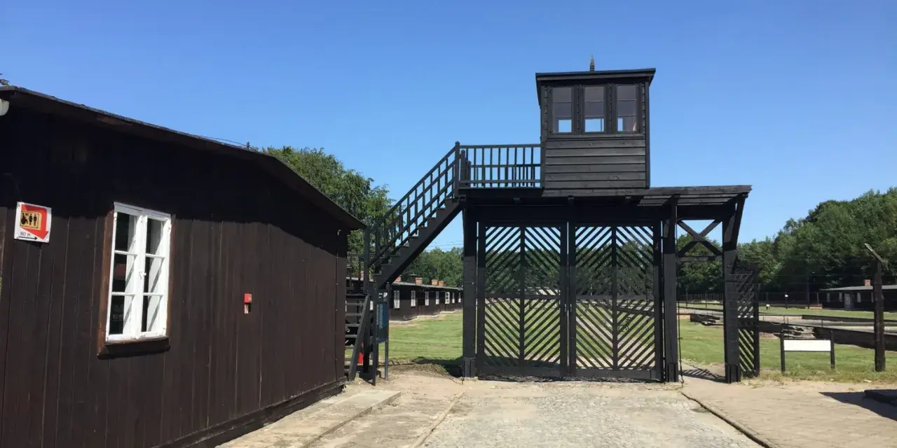 Brama i wieża strażnicza na terenie Muzeum Stutthof. Drewniane budynki i brukowany chodnik.