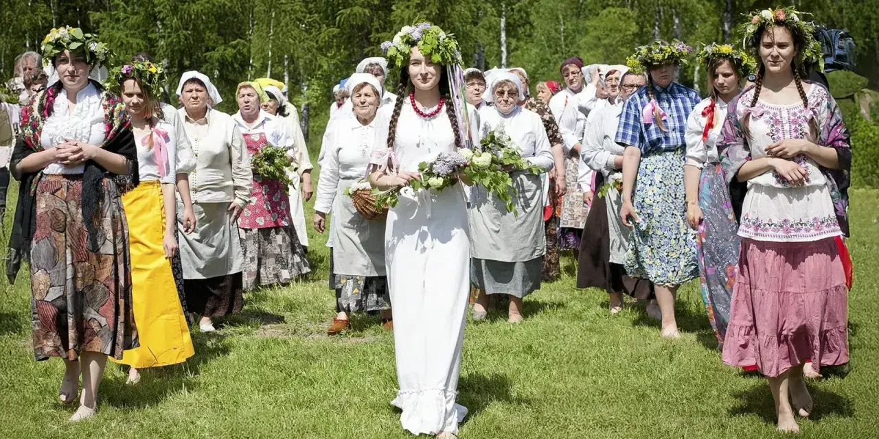 Grupa kobiet wiankach na głowach i tradycyjnych strojach idzie przez łąkę. Obraz nawiązuje do święta zielone świątki.