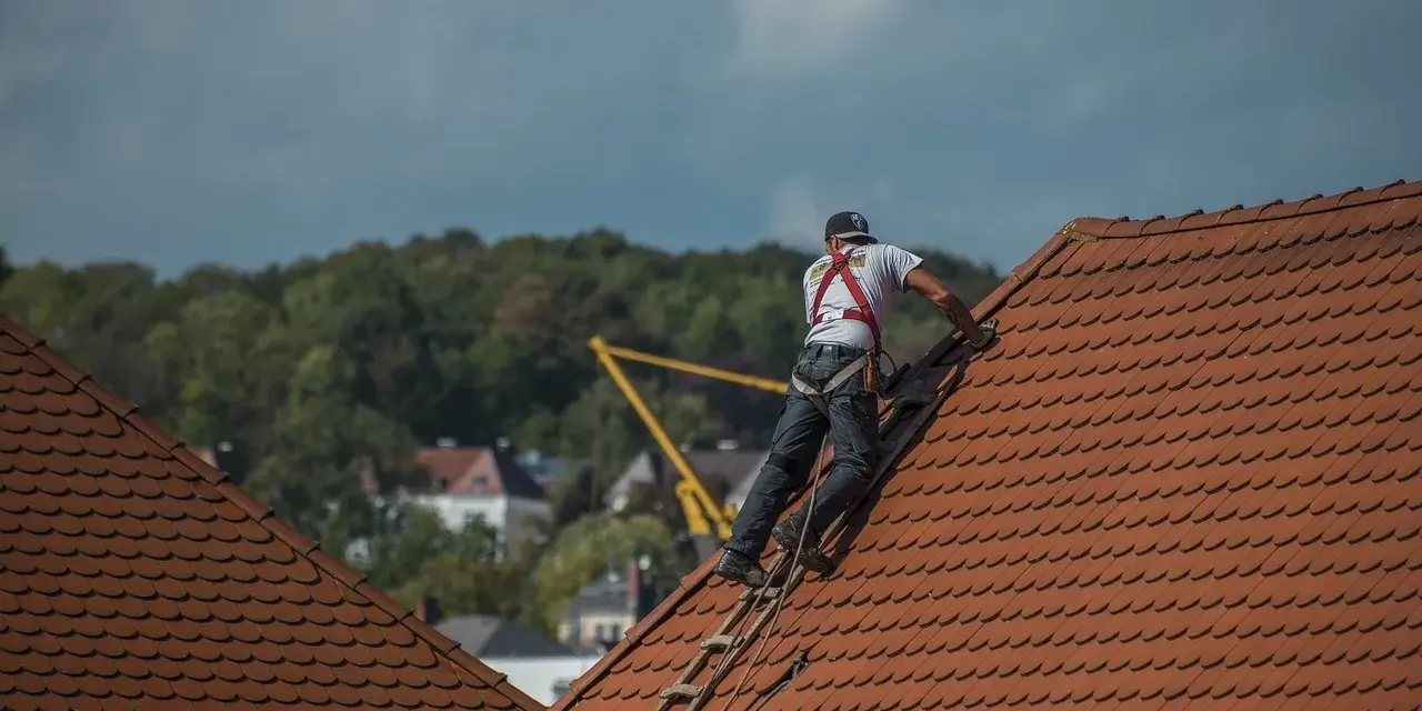 Robotnik w kasku i szelkach bezpieczeństwa na dachu, pracujący przy dachówce.