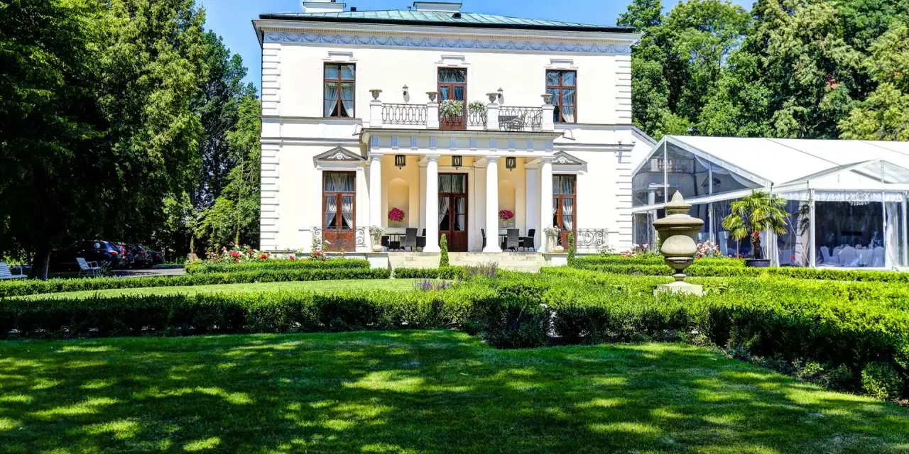 Elegancki pałac Rozalin z kolumnowym portykiem i balkonem, otoczony zielonym ogrodem i nowoczesnym namiotem bankietowym.