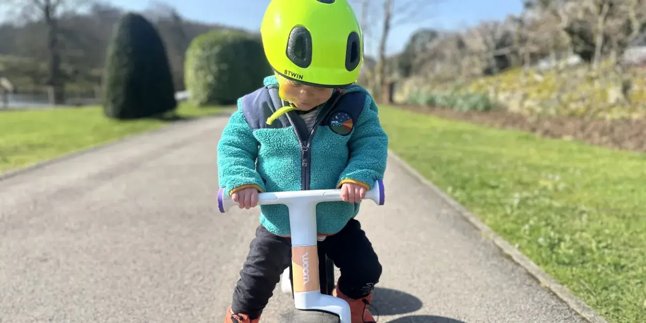 A child in a bright green helmet and teal jacket rides a woom wow balance bike on a sunny path.