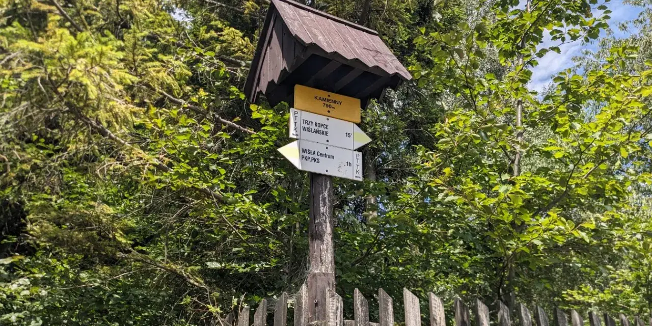 Znak szlaku PTTK w lesie, wskazujący drogę na Trzy Kopce Wiślańskie i do Wisły. W tle zieleń drzew i drewniany płot.
