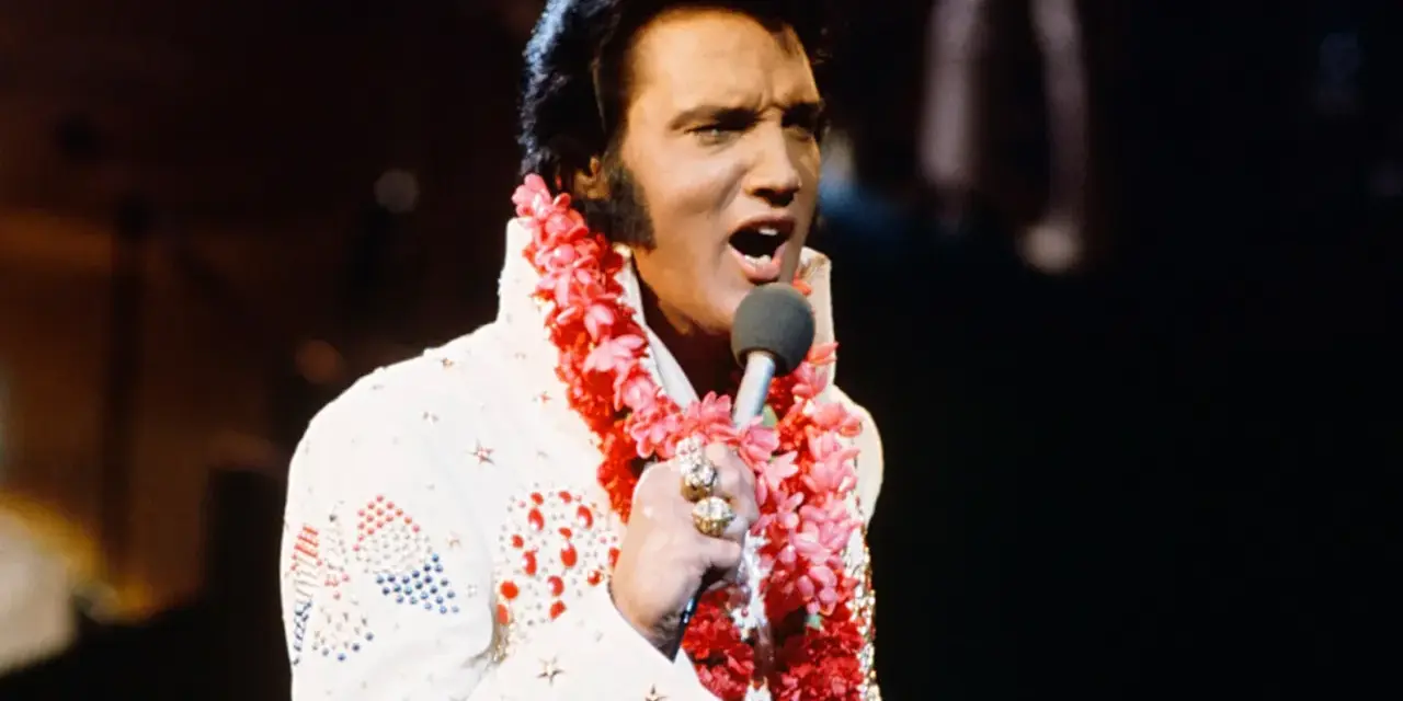 Elvis Presley singt in Hawaii, trägt einen weißen Anzug mit Blumenkette und Mikrofon.