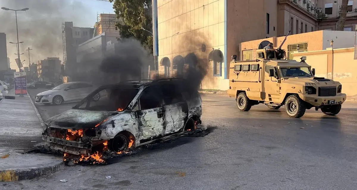 Płonący samochód i pojazd opancerzony na ulicy. Obraz sugeruje, czy Libia jest bezpieczna, pokazując chaos i obecność wojska.