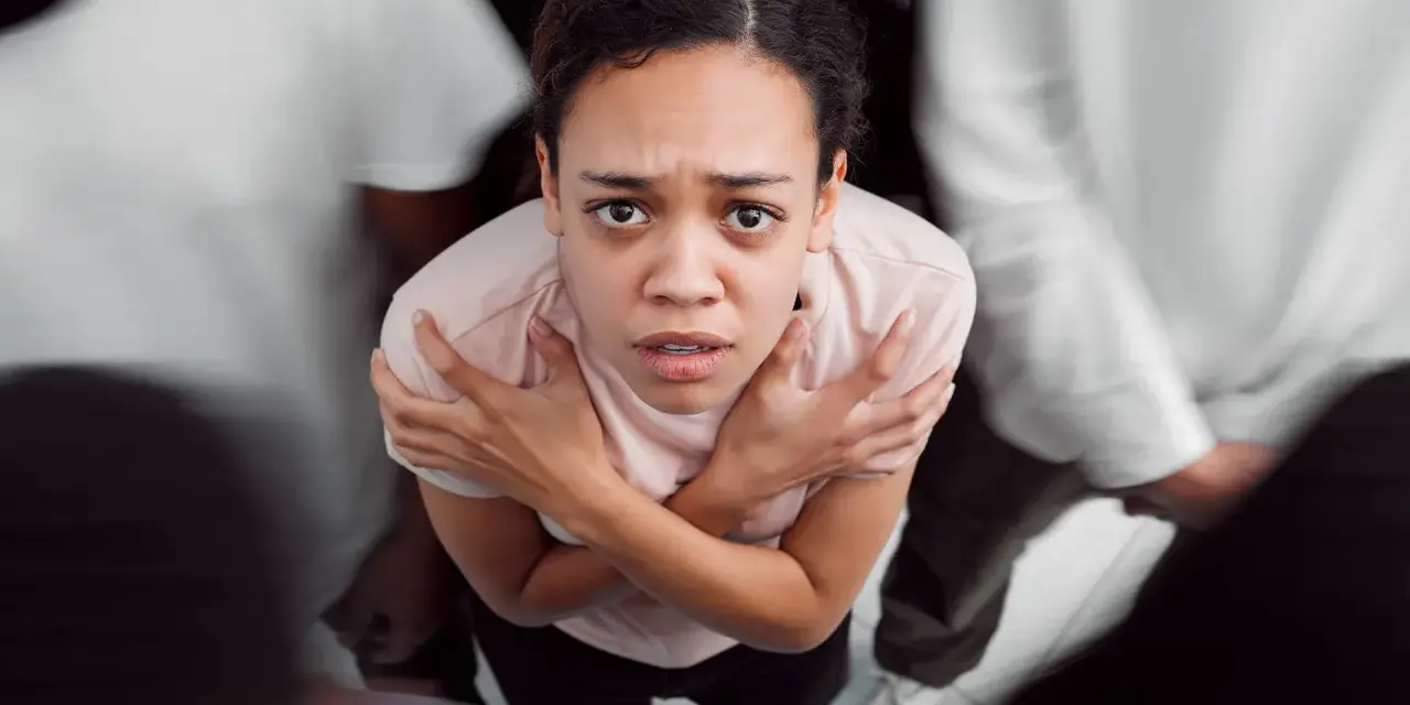 Donna spaventata, circondata da figure sfocate, con le braccia incrociate sul petto. Potrebbe rappresentare i disturbi psicosomatici, un elenco di sintomi fisici legati allo stress.
