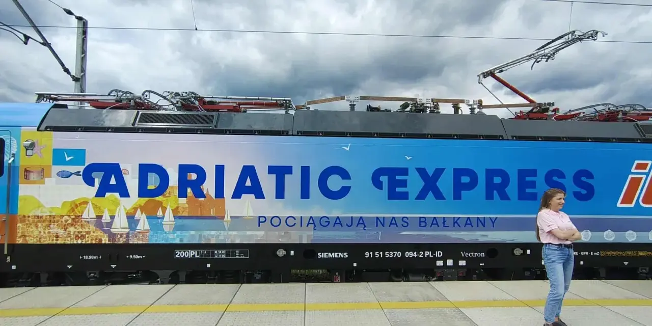 Kobieta stoi obok lokomotywy Adriatic Express z widokiem na morze i żaglówki.