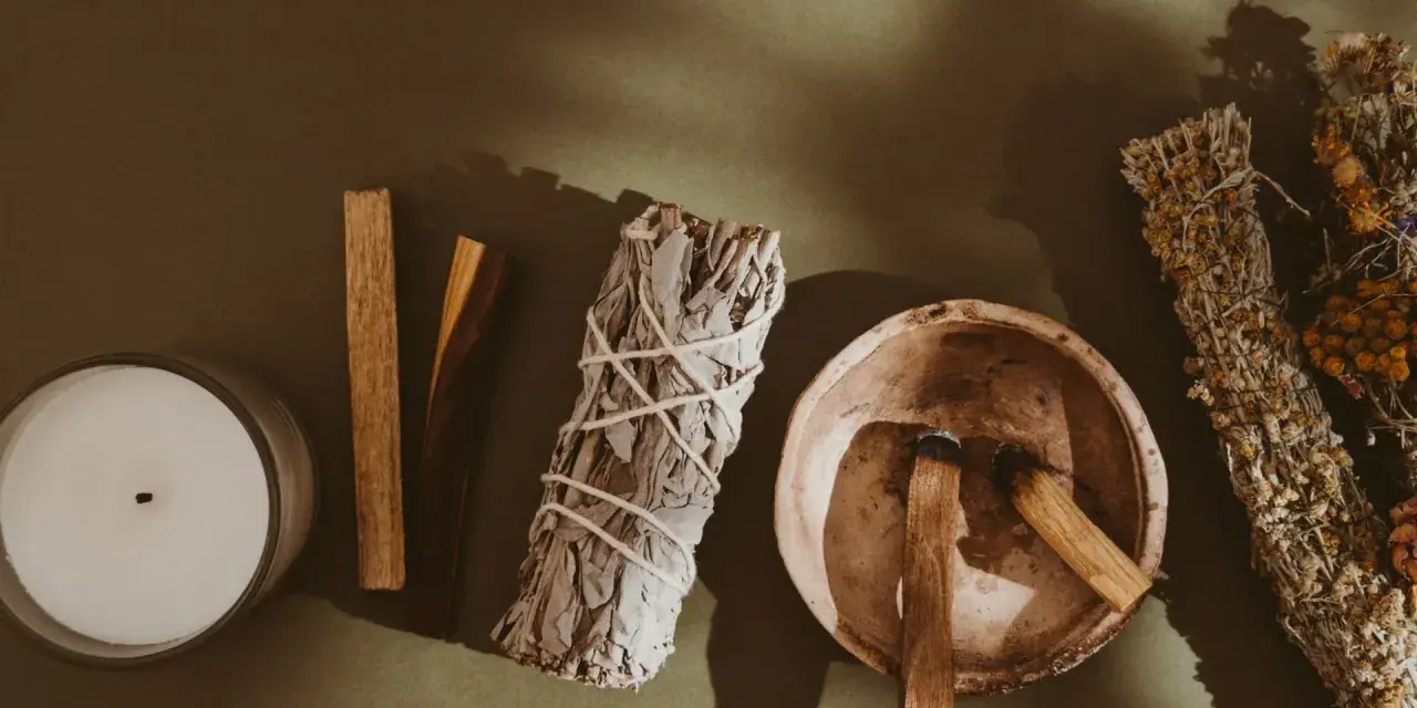 Palo santo kadzidło właściwości: świeca, biała szałwia, miseczka z drewienkami palo santo i bukiety ziół.