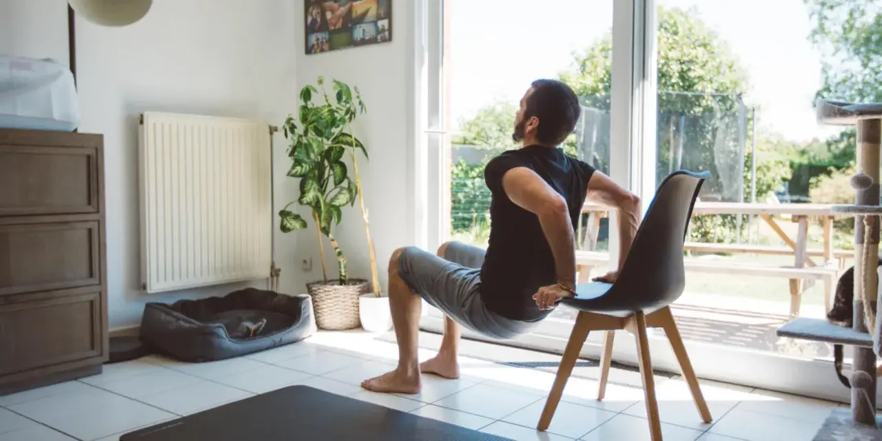 Un homme fait des dips sur une chaise, une idée de quel sport faire à la maison. Un tapis de yoga est au sol.