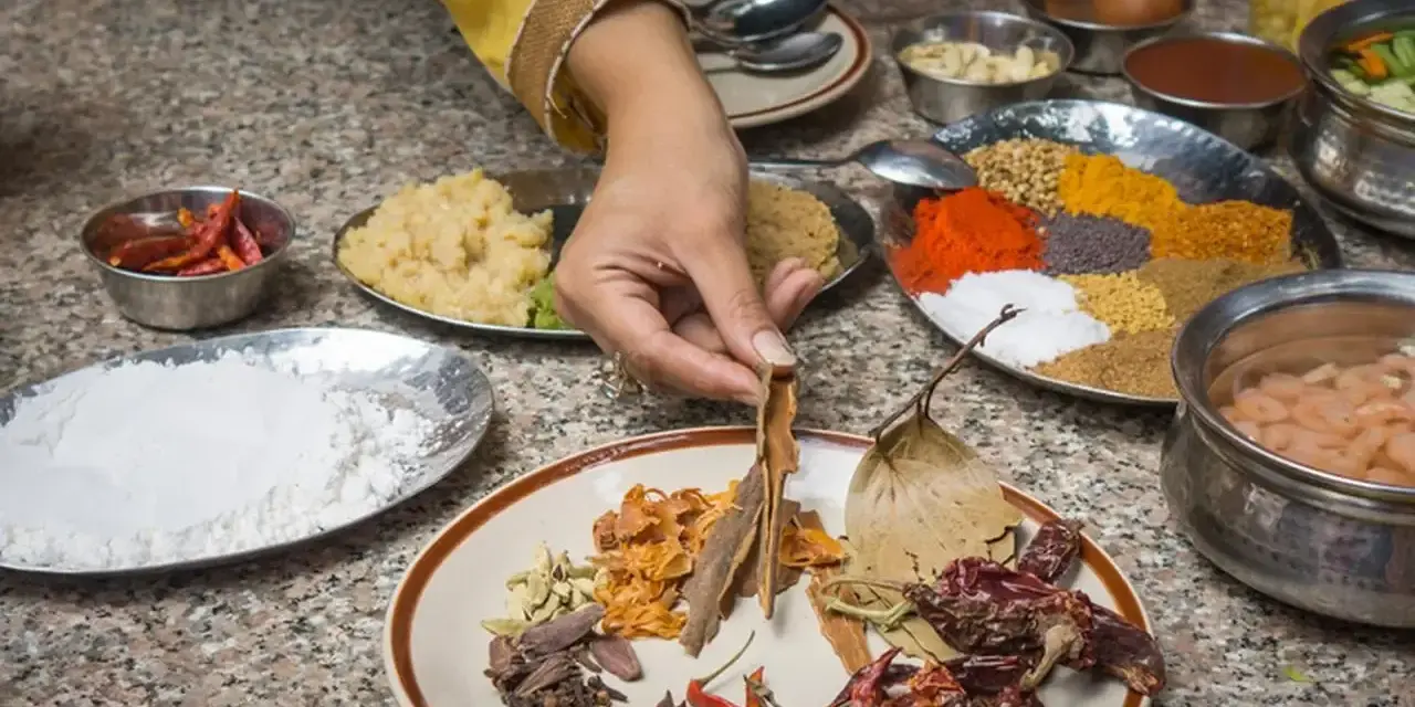 Ręka wybiera cynamon z talerza przypraw, przygotowując autentyczną kuchnię nepalską. Widać chili, mąkę, pastę imbirową i kolorowe przyprawy.