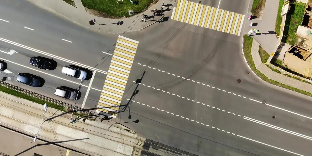 Vista aerea di un incrocio con strisce pedonali bianche e gialle. Auto in attesa nella corsia di preselezione, tram sui binari sottostanti.