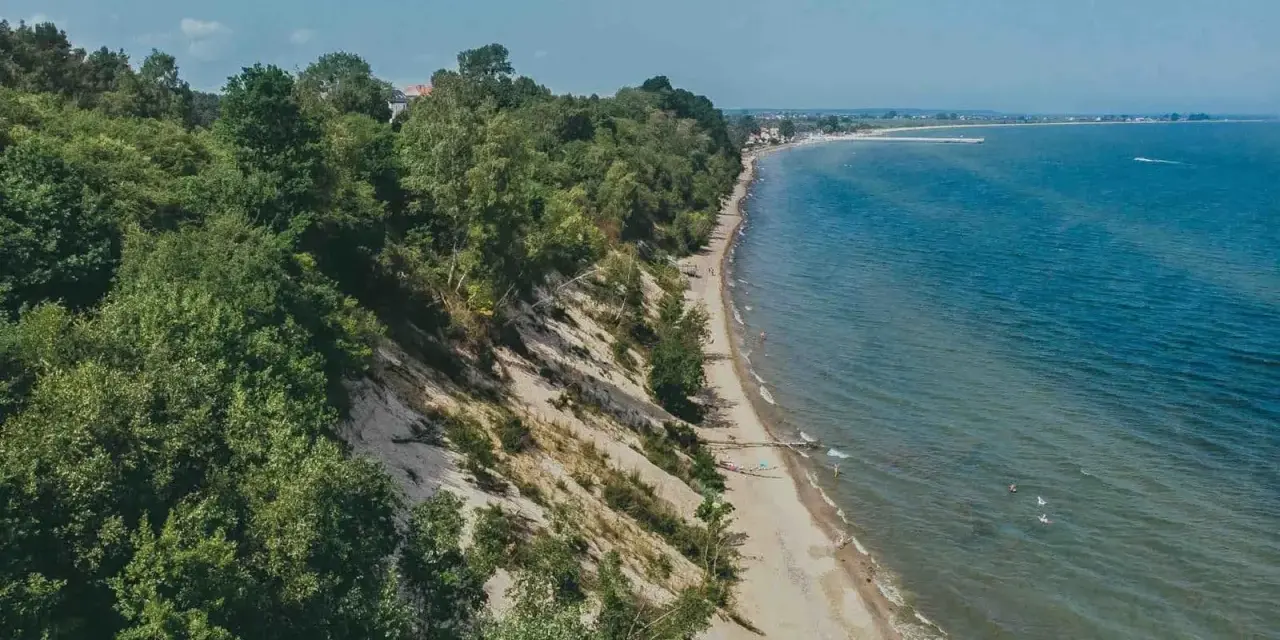 Mechelinki klif porośnięty drzewami, z piaszczystą plażą i turkusowym morzem. W oddali widać zabudowania i falochron.