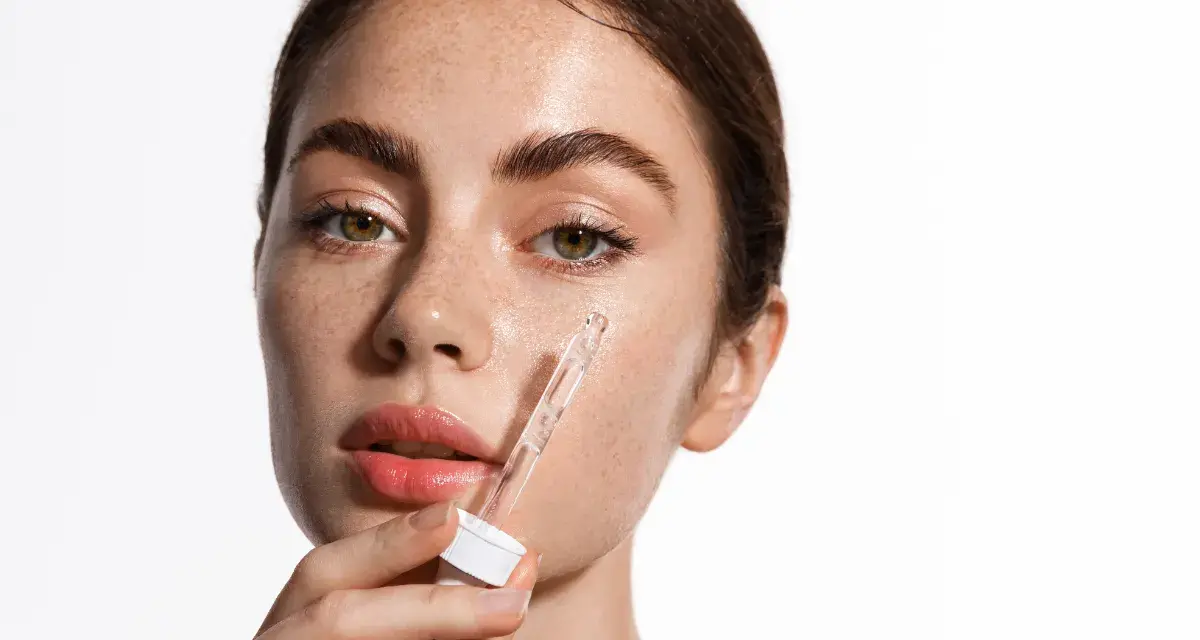 Kobieta aplikuje serum z pipetki na policzek. Skóra pełna piegów, nawilżona, z widocznym blaskiem. Kluczowe składniki: retinol i ceramidy.