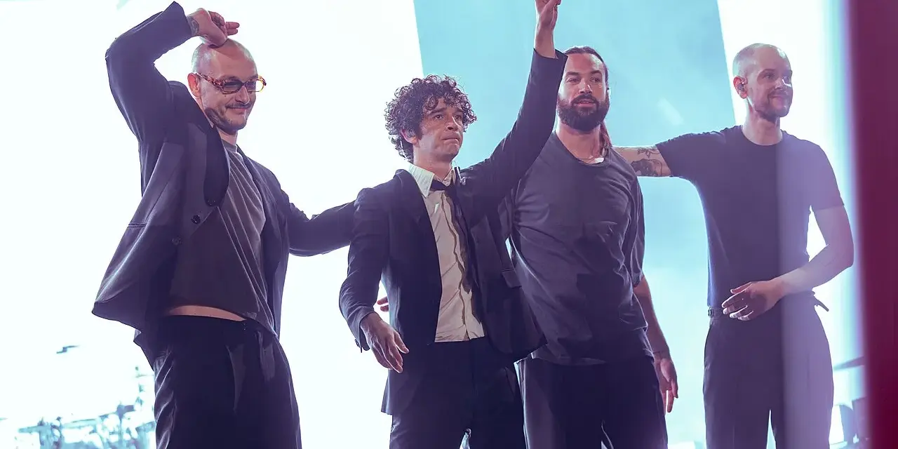 Matty Healy i zespół The 1975 na scenie. Muzycy pozdrawiają publiczność, jeden z nich unosi rękę w geście pokoju.