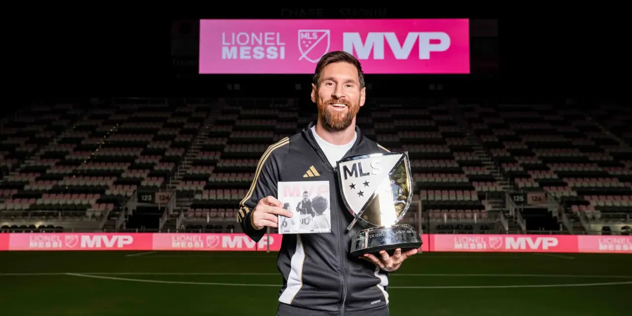 Lionel Messi sonríe en un estadio, sosteniendo un trofeo MLS MVP y un libro. El letrero del estadio dice "LIONEL MESSI MVP".