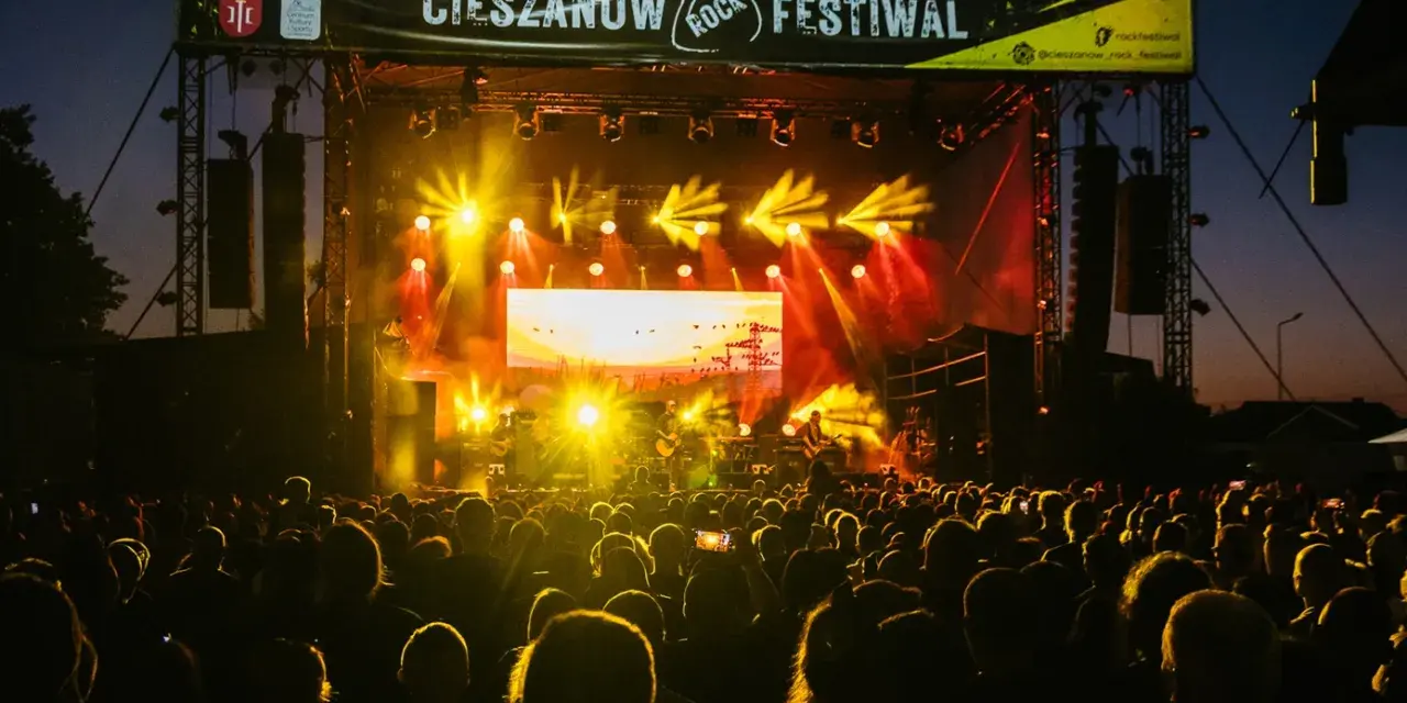 Tłumy bawią się na Cieszanów Rock Festiwal. Jasne światła sceny oświetlają zespół grający na tle zachodzącego słońca.