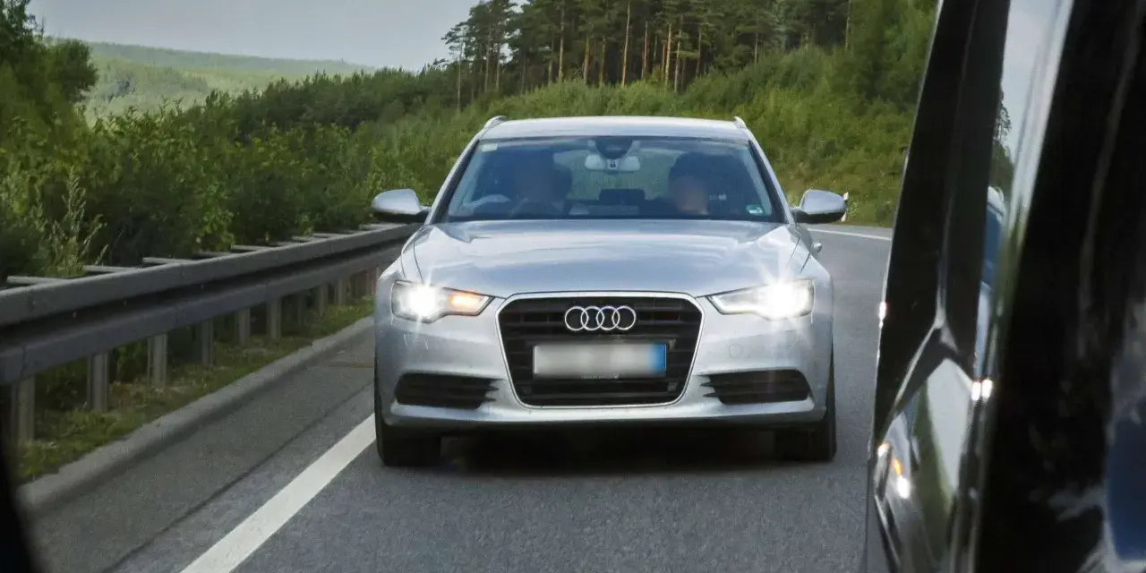 Silberner Audi auf der Autobahn, blendet mit Lichthupe. Vorbei an grünen Wäldern und einer Leitplanke.