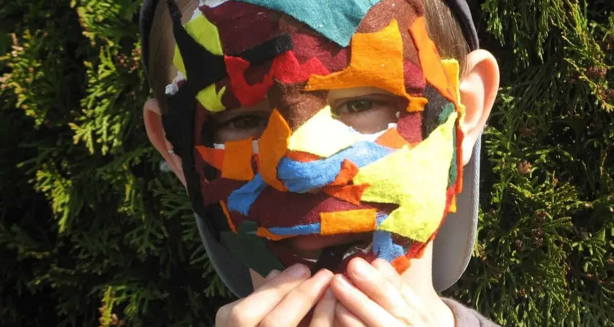 Dziecko w kolorowej masce z filcu, symbolizującej maskowanie autyzm. Dłonie przy ustach.