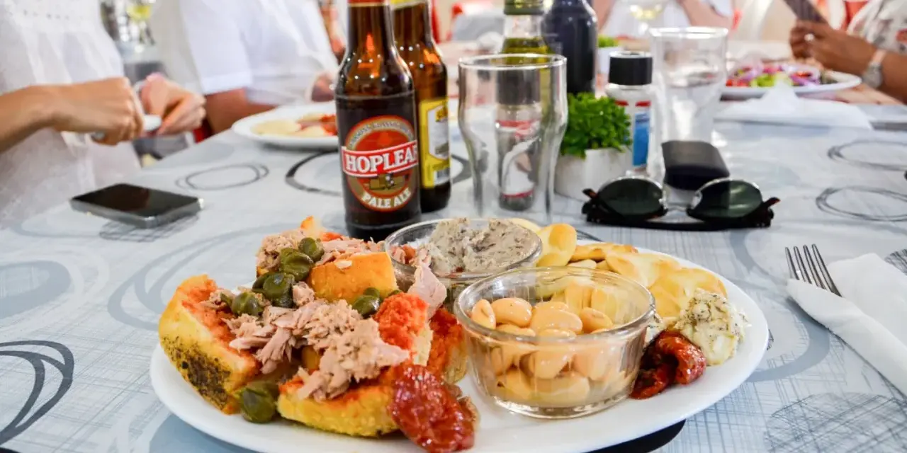 Malta jedzenie: talerz pełen smakołyków z tuńczykiem, fasolą i oliwkami, obok piwo i ludzie przy stole.