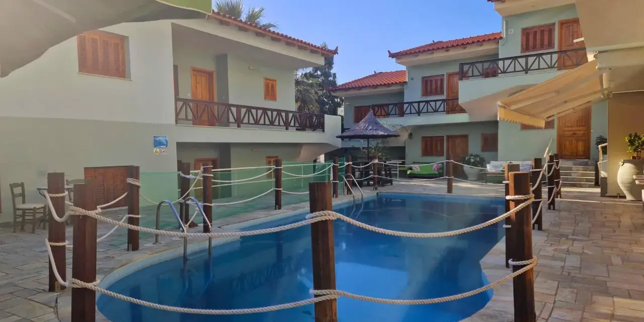 Piscine bleue entourée de cordes et de poteaux en bois, avec des bâtiments de style colonie en Grèce en arrière-plan.
