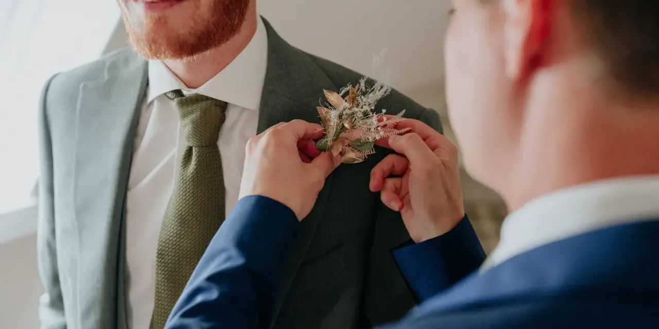 Un homme ajuste une boutonnière sur le costume d'un autre. Divers types de boutonnières existent, celle-ci est composée de fleurs séchées et de feuillages dorés.