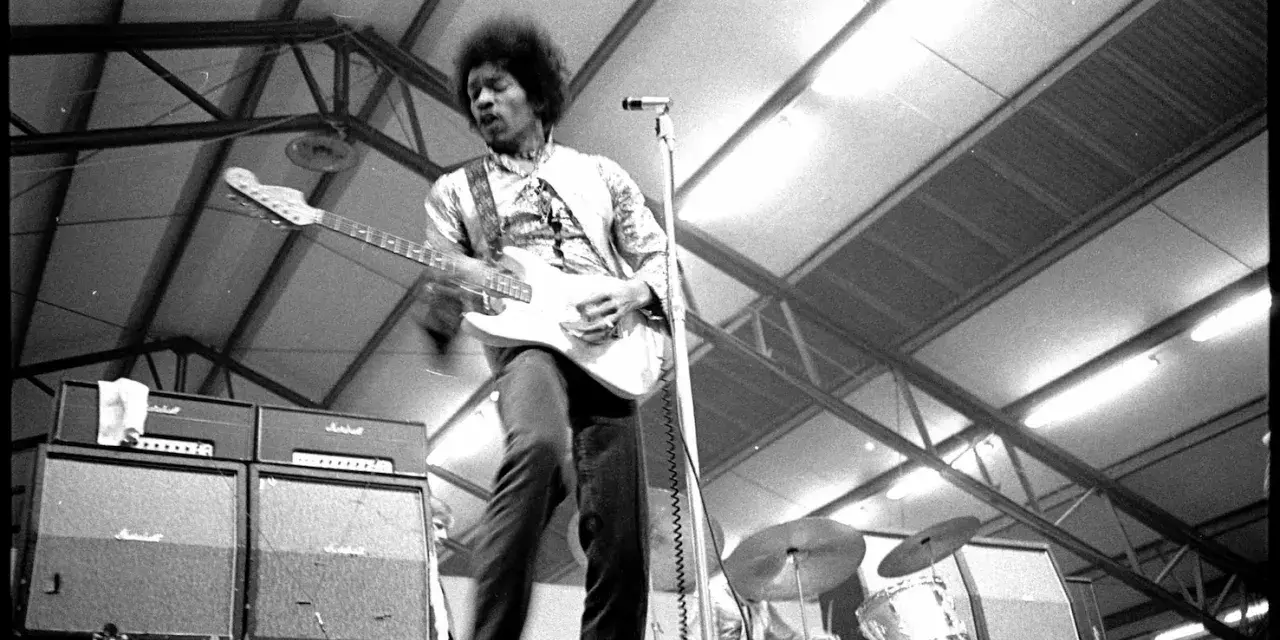 Jimi Hendrix, icône du jimi hendrix style de musique, électrise la scène avec sa guitare blanche. Amplis Marshall et batterie en arrière-plan.