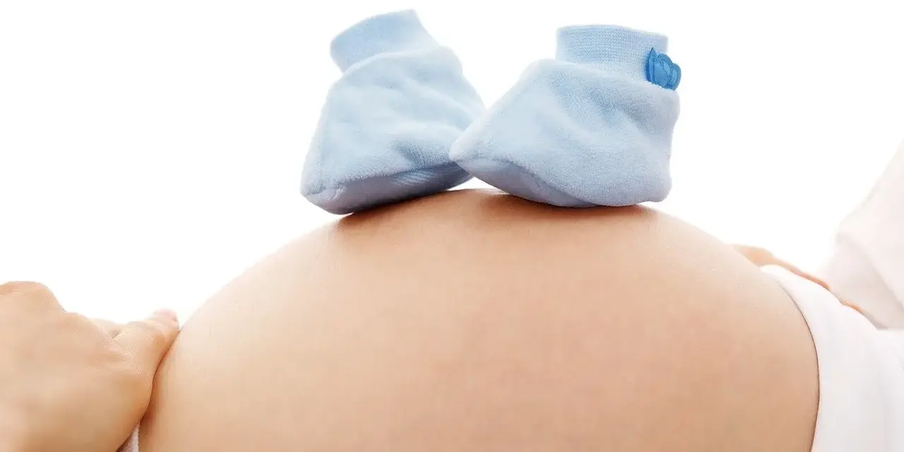 Cofnięcie wypowiedzenia umowy o pracę z powodu ciąży wzór. Dłoń kobiety na brzuchu ciążowym, na którym leżą niebieskie buciki dla niemowlaka.
