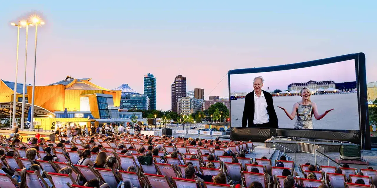 Ein lauer Sommerabend im Freiluftkino Charlottenburg. Menschen sitzen in Liegestühlen und schauen auf eine Leinwand, auf der ein Film mit einem älteren Mann und einer jungen Frau läuft. Im Hintergrund die Berliner Skyline.