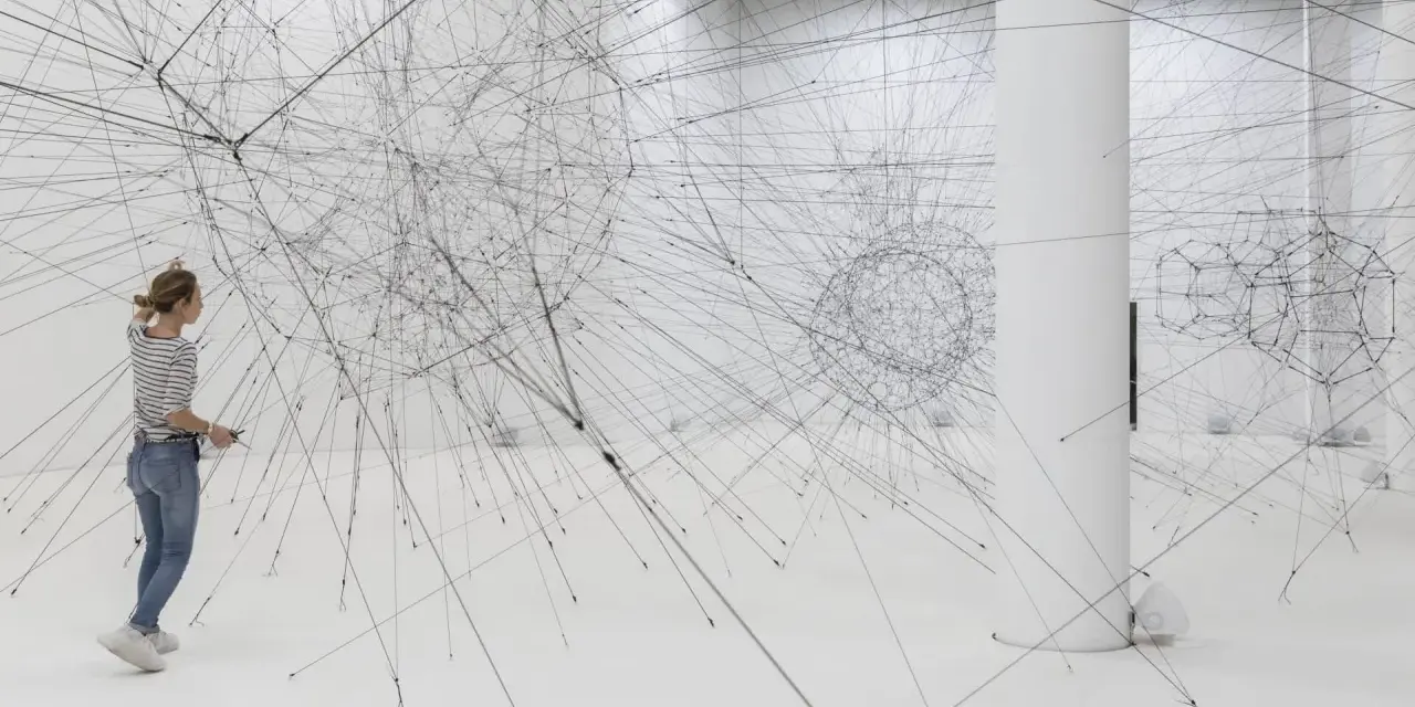 Une femme admire une installation de Tomas Saraceno, un réseau complexe de fils noirs tendus dans un espace blanc.