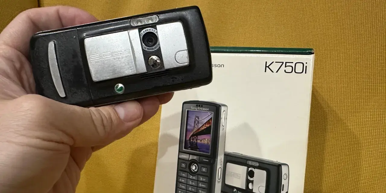 Dłoń trzyma czarny telefon Sony Ericsson K750i, obok stoi pudełko z tym modelem.