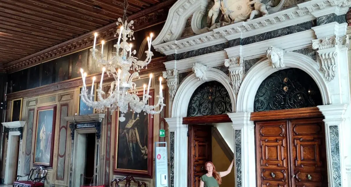 Una donna in un abito verde posa davanti a un portale decorato in una casa museo a Venezia, con un lampadario di cristallo e dipinti alle pareti.