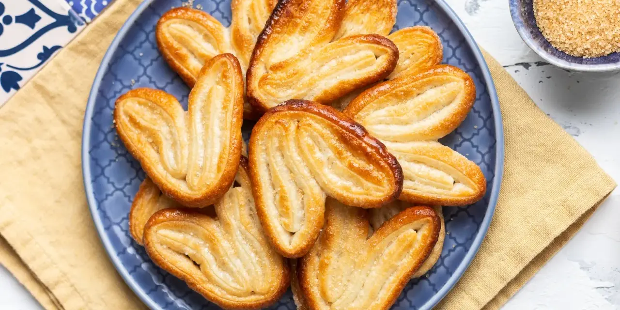 Un piatto blu pieno di dolci con pasta sfoglia dorati e croccanti, cosparsi di zucchero.