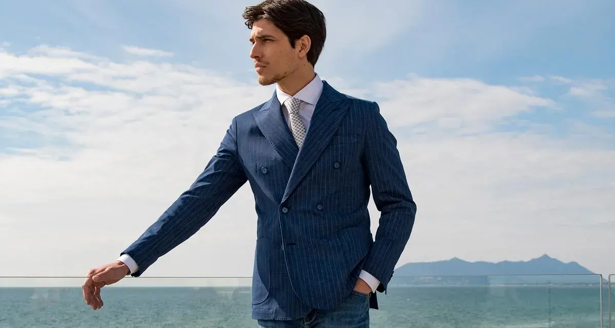 Uomo con un elegante outfit uomo estate: giacca doppiopetto blu a righe, camicia bianca, cravatta e jeans. Vista mare.