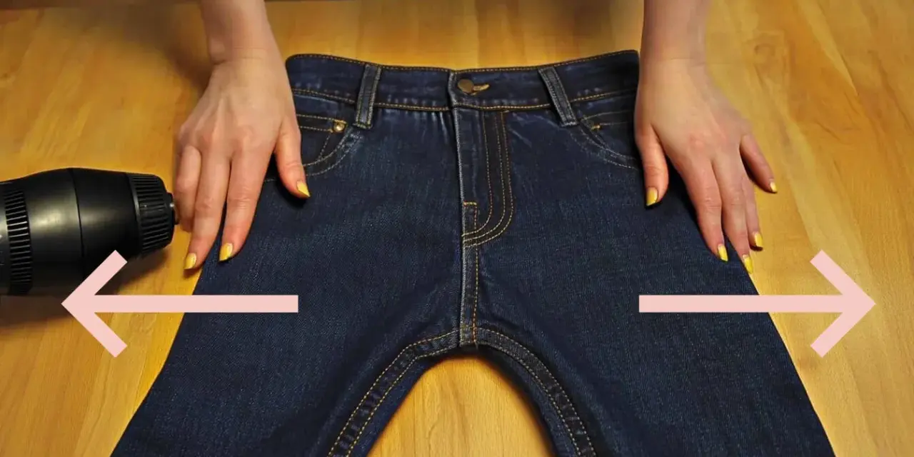 Manos sujetan pantalones vaqueros oscuros sobre mesa de madera. Secador de pelo apunta a la tela. Aprende como estirar pantalones vaqueros.