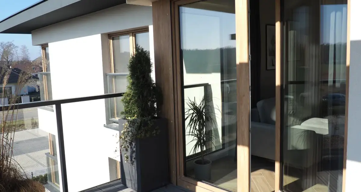 Nowoczesny balkon z drewnianymi, podwójnymi drzwiami balkonowymi. Okna dzielone poziomo wpuszczają dużo światła.