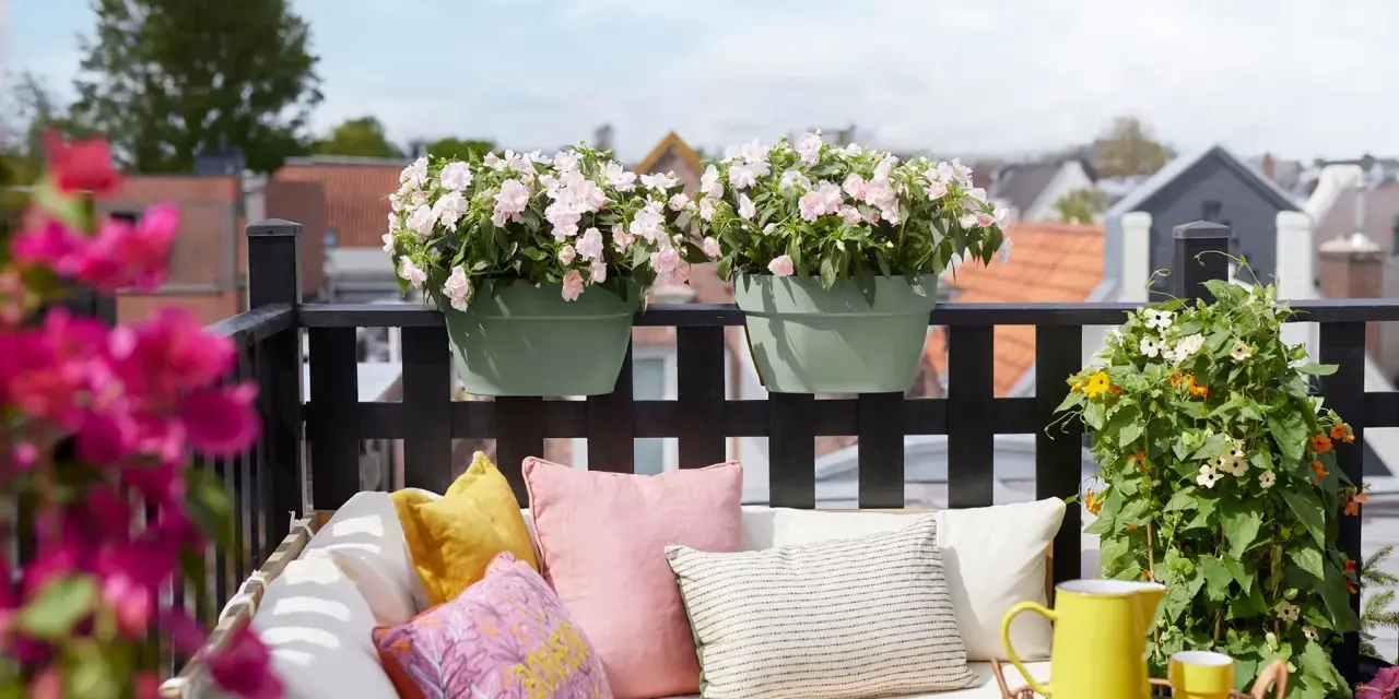 Ein gemütlicher Balkon mit bunten Kissen, gelber Kanne und blühenden sommerblumen.