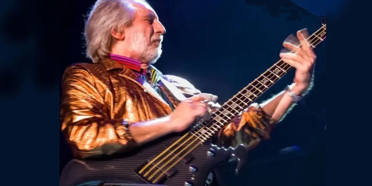 Ein Mann mit grauem Haar und Bart spielt Bassgitarre. Er trägt eine glänzende goldene Jacke und bunte Krawatte. Er ist der beste Bassist der Welt.