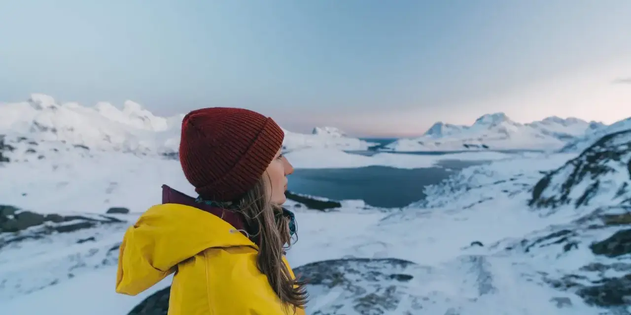 Kvinna i gul jacka och röd mössa njuter av utsikten över snöklädda berg och hav. Perfekt för sportlov i Norge.