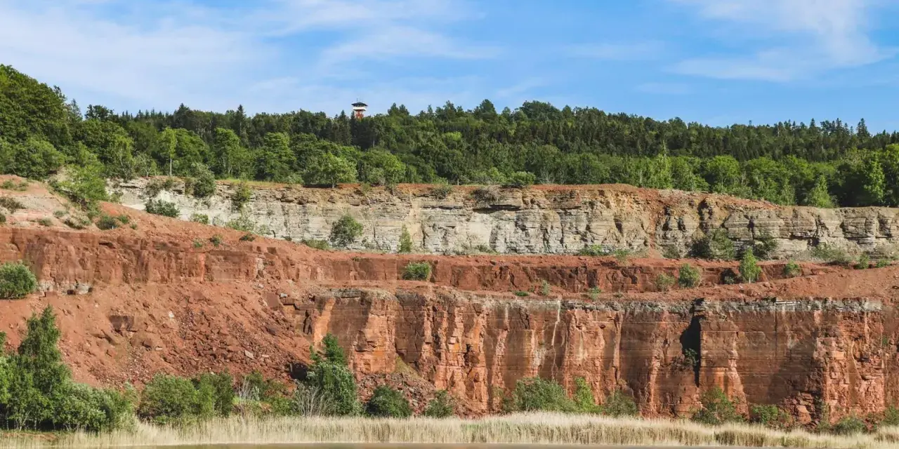 En sjö speglar Sveriges Grand Canyon, en imponerande klippformation med lager av röd och grå sten, krönt av en grönskande skog.