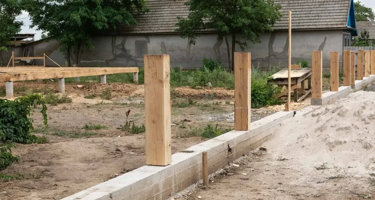 Budowa fundamentu pod ogrodzenie: betonowy murek, drewniane słupki i piasek.