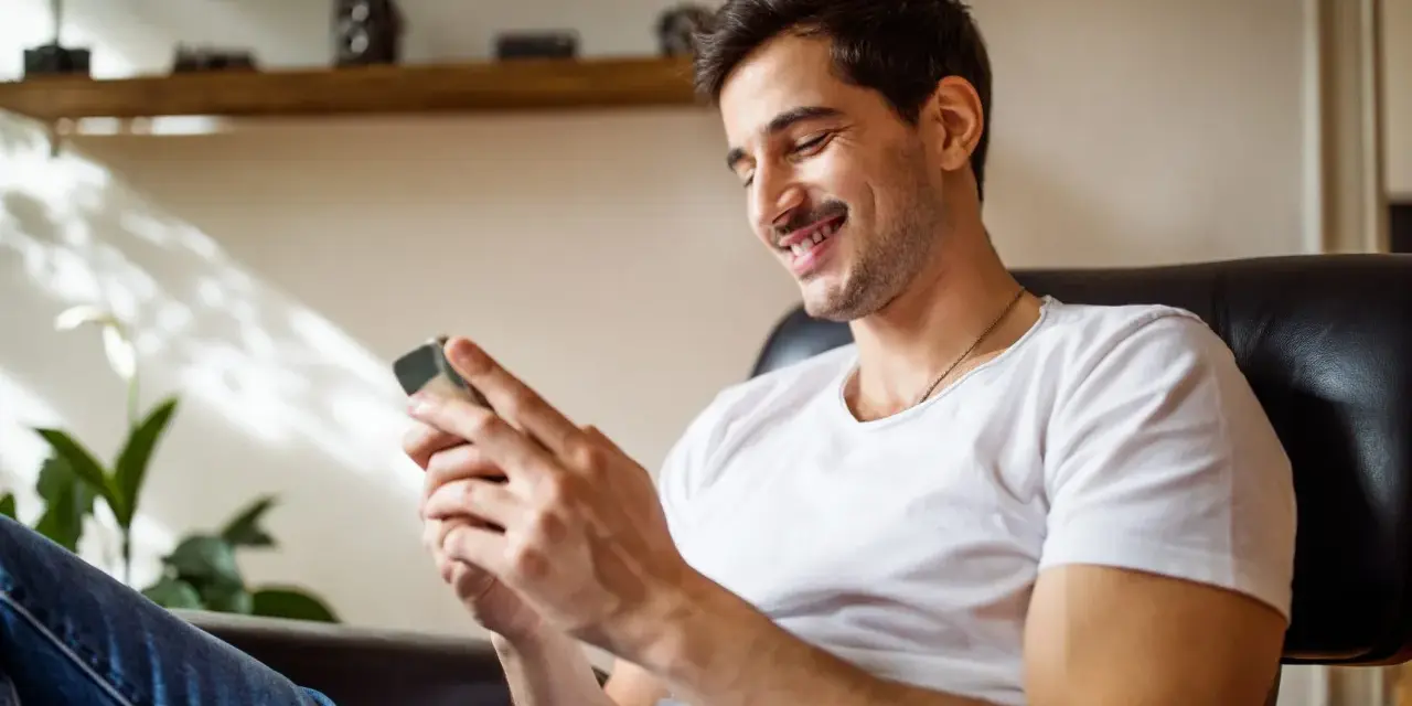 Uśmiechnięty mężczyzna w białej koszulce siedzi w fotelu i patrzy na telefon.